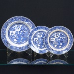FAIANÇA INGLESA, três (3) pratos: 1 fundo com bicado na borda; 2 de sobremesa, ambos com trincados, ornamentação dita WILLON (Segundo a lenda chinesa: Um certo Mandarim tinha uma leal e devotada filha, possuidora de rara beleza. Certo dia, em que descobre que ela estava apaixonada por um carteiro que lhe fazia a corte, e que era por ela correspondida, expulsa a filha de casa e roga-lhe uma praga: que ao ser trocado o primeiro beijo pelo casal, eles se transformariam em dois pombos), também conhecida como pombinhos, manufatura WOOD & SONS Ltd. (fundada em 1865, funcionou na Potteries New Wharf em Trent e mais tarde na Pottery Stanley em Burlem, fechou em 2005), tonalidade azul sobre fundo branco, medindo respectivamente 23 cm e 18 cm diâmetro, possuem o craquelê característico do envelhecimento de toda a faiança, sinais de uso, vide foto.