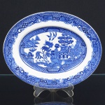 FAIANÇA INGLESA, uma (1) travessa oval, ornamentação dita WILLON (Segundo a lenda chinesa: Um certo Mandarim tinha uma leal e devotada filha, possuidora de rara beleza. Certo dia, em que descobre que ela estava apaixonada por um carteiro que lhe fazia a corte, e que era por ela correspondida, expulsa a filha de casa e roga-lhe uma praga: que ao ser trocado o primeiro beijo pelo casal, eles se transformariam em dois pombos), também conhecida como pombinhos, manufatura WOOD & SONS Ltd. (fundada em 1865, funcionou na Potteries New Wharf em Trent e mais tarde na Pottery Stanley em Burlem, fechou em 2005), tonalidade azul sobre fundo branco, medindo 26 x 20 cm, possui o craquelê característico do envelhecimento de toda a faiança, sinais de uso, vide foto.