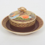 FAIANÇA JAPONESA, uma (1) manteigueira com presentoir acoplado e tampa, ornamentação policromada representando dignatários encimado pelo simbolo de Satsuma, estilo SHOZA (criado por Shoza na região de Kutani, em finais do período Edo no século XIX, com sua principal característica cores berrantes e generosos acabamentos a ouro, quando morreu em 1883 já tinha mais de 300 aprendizes o que fez com que o estilo perdurasse até a atualidade), pega da tampa relevada em cabeça de dragão celestial, manufatura J.H. dentro de um losango e a legenda Made in Japan, medindo 20 cm altura, possui manchas e o craquelê característico do envelhecimento de toda a faiança, medindo 13 cm diâmetro, sinais de uso, vide foto.