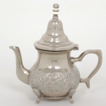 METAL PRATEADO, um (1) bule oriental, bojo com ornamentação relevada no padrão Império Otomano, pés em concha terminando em cabriole, adorno da extremidade da tampa em polímero na tonalidade branca, marca de origem ou manufatura na base em Árabe, medindo 19 cm altura, sinais de uso, vide foto.