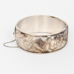 Joia, Prata. Bracelete oval confeccionado em prata portuguesa, decoração relevada com padrão de flores e folhagens, obtidas através das técnicas do repuxado e cinzelado, fecho tipo gaveta com corrente pega ladrão. Apresenta marca de garantia da prata do toque 0,833, usada na contrastaria do Porto, da joalheria Rosas de Portugal e punção de ourives. Medidas: Alt.: 2.5 x Comp.: 7 x Larg.: 6.5 cm. Peso: 37 gramas.