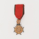 Cruz de Combate  1ª Classe  Força Expedicionária Brasileira (FEB). Executada em prata com vermeil. Insígnia constituída por cruz pátea de quatro braços com extremidades arredondadas, sobreposta a feixe de raios. Ao centro, medalhão circular com estrelas em relevo circundadas por coroa. Suspensa por elo intermediário com emblema contendo as iniciais FEB. Reverso com inscrição: ESTADOS UNIDOS DO BRASIL  CRUZ DE COMBATE  1ª CLASSE. Fixação por argola a fita de gorgorão vermelha com frisos verdes laterais. Executada em prata. Medidas: Medalha: Diam.: 1,5 cm. Comp.: 5 cm.