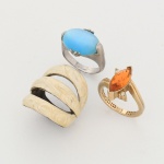 Três(03) itens: a) Anel confeccionado em metal dourado, apresenta pedra central de coloração laranja, lapidação navete facetada, cravada por garras. Ladeando a pedra central, duas gemas incolores de lapidação facetada, cravadas individualmente. Medida: Diam.: 2 cm. b)  Anel confeccionado em metal, composto por aro de seção circular, alargando-se na parte superior. Apresenta pedra azul em formato oval confeccionada em material sintético, fixada por garras laterais e complementada por elementos metálicos superiores e inferiores. Estrutura com galeria vazada sob a pedra. Medida: Diam.: 2 cm. c) Anel confeccionado em metal, constituído por estrutura composta por três aros paralelos interligados, formando corpo único, faces externas revestidas em esmalte de tonalidade marfim, com padrão marmorizado, construção vazada entre os aros, formando intervalos longitudinais. Medidas: Comp.: 3 cm. Diâmetro: 2 cm.