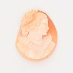 Camafeu esculpido em concha bicolor, de formato oval, apresenta busto feminino em perfil voltado à direita, com cabelo ondulado preso por diadema, brinco, colar e indumentária com pregueados, esculpida em relevo aproveitando o contraste natural das camadas da concha.Verso liso e polido. Apresenta trincado, e quebradura com fragmento lateral destacado. Demanda restauro. Medidas: Alt.: 5 x Comp.: 4 cm.