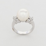 Anel em metal prateado com pérola de imitação e pedras sintéticas. Elemento central constituído por esfera com revestimento nacarado, fixada por pino. Laterais compostas por pedras incolores de lapidação brilhante, cravadas em garras, dispostas em agrupamentos simétricos. Medida: Diam.: 2 cm.