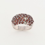 JOIA. PRATA. Anel confeccionado em prata, frente larga inteiramente pavimentada por granadas de coloração avermelhada(apresenta duas perdas), de lapidação facetada, cravadas em garras, dispostas em fileiras horizontais contíguas. Galeria interna vazada com perfurações correspondentes às cravações. Aro de secção abaulada. Medida: Diam.: 2 cm. Peso: 7,7 gramas.