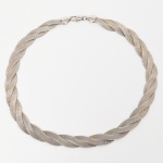 Colar confeccionado em metal prateado, composto por três fitas de malha flexível entrelaçadas em padrão helicoidal, formando estrutura trançada contínua, extremidades com terminais cônicos lisos, e fecho tipo lagosta com argola de união. Apresenta pequenos desgastes e marcas de uso, visíveis nas fotografias. Medida aberta: 45 cm.