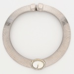 Colar confeccionado em metal prateado, composto por estrutura tubular articulada, formada por segmentos metálicos justapostos com acabamento canelado transversal, conferindo flexibilidade ao conjunto, fecho tipo caixa com lingueta articulada e encaixe por pressão.Na parte frontal, aplicação central oval composta por pedra incolor translúcida em material sintética, montada em engaste liso, circundando o cabochão, moldura oval guarnecida por fileira contínua de pequenas pedras incolores facetadas, cravadas individualmente. Apresenta pequenos desgastes de uso, vide foto. Medida fechada: Diam.: 15 cm.