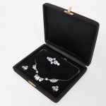 Conjunto confeccionado em metal prateado, ornado com pedras lapidadas em tonalidade lilás  provável década de 1940/1950, composto por colar, broche e par de brincos, acondicionado em estojo. Colar estruturado em corrente de malha fina com fecho tipo mola. Parte frontal articulada composta por elementos geométricos dispostos simetricamente, apresentando pedras centrais em lapidação octogonal (tipo esmeralda), intercaladas por pedras circulares em lapidação brilhante e elementos retangulares em lapidação baguete. Montagem em garras e engastes vazados posteriores. Broche de composição linear simétrica, com duas pedras octogonais centrais ladeadas por pedras circulares. Sistema de fixação por alfinete horizontal com trava rotativa. Brincos em composição triangular, com três pedras circulares em lapidação brilhante e duas pedras retangulares em lapidação baguete, montadas em garras. Sistema de fixação por fecho de pressão tipo clip. Estrutura em metal de tonalidade prateada. Pedras translúcidas em tonalidade lilás, possivelmente vidro lapidado ou cristal sintético, compatíveis com produção industrial do período. Estojo revestido externamente em material têxtil e internamente em veludo. Apresenta desgaste no banho de prata, marcas de uso e do tempo, vide foto. Medidas: Colar(medida aberta): 40 cm; Broche: Comp.: 4,5 cm; Brincos: Alt.: 1,5 x Comp.: 1,5 cm.