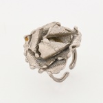 Anel confeccionado em metal prateado, composição escultórica representando flor, estruturada por pétalas sobrepostas com bordas onduladas, superfície com acabamento texturizado, elemento central composto por esfera metálica em tonalidade dourada, parcialmente envolvida pelas pétalas internas. Aro duplo, aberto na parte inferior, seção circular, fixado à base da estrutura floral. Apresenta oxidação e marcas de uso, vide foto. Medidas: Aro: Diam.: 2,5 cm. Flor: 3,5 cm.