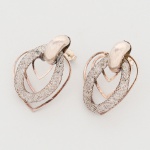 Par de brincos em metal prateado, constituídos por elemento superior ovalado, de superfície polida, ao qual se articulam três módulos sobrepostos em formato de coração estilizado. O módulo externo apresenta contorno vazado em fio metálico; o módulo intermediário repete o desenho em escala reduzida; o módulo inferior é composto por malha tubular flexível, tipo trama entrelaçada (malha trançada metálica), conformada em desenho cardíaco. Estrutura articulada por meio de eixo metálico posterior. Sistema de fixação por pino e fecho de pressão (tarraxa tipo borboleta). Medidas: Alt.: 3,5 x Comp.: 2,5 cm.