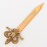 Marcador de livros em metal dourado com motivo heráldico, acabamento dourado, composto por lâmina plana de seção retangular, terminando em ponta ogival chanfrada, extremidade superior apresenta elemento aplicado em relevo representando flor-de-lis coroada, ladeada por volutas estilizadas e travessas horizontais ornamentais. A composição remete a motivo heráldico de tradição francesa. Apresenta desgaste no douramento. Medida: Comp.: 8 cm.