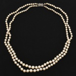 JOIA. Colar de duas voltas com pérolas e fecho em prata, composto por duas voltas de pérolas cultivadas, de tonalidade creme, enfiadas em fio contínuo, com nós de separação visíveis entre os elementos. Pérolas de formato predominantemente circular, com variação dimensional discreta ao longo dos fios. Fecho retangular confeccionado em prata, estruturado em duas placas articuladas, decorado com pedrarias incolores de lapidação brilhante, dispostas em composição geométrica com motivos losangulares e elemento circular central. Sistema de fecho tipo caixa com lingueta de encaixe e dispositivo de segurança lateral. Medida: Comp.: 25 cm.