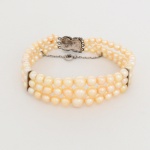 JOIA. Pulseira Art Déco composta de quatro fileiras de pérolas cultivadas, estilo Art Déco, de formato predominantemente esférico a levemente ovalado, tonalidade creme a levemente dourada, com brilho nacarado, encontram-se enfiadas com separação individual por nós, intercalados por placa retangular em metal prateado, organizando estruturalmente as fileiras.Fecho confeccionado em prata formado por duas placas articuladas, decoradas com pedrarias incolores de lapidação brilhante, dispostas em composição geométrica com motivos losangulares e elemento circular central, característicos do repertório formal do Art Déco.Sistema de fecho tipo caixa com lingueta de encaixe, acompanhado de corrente de segurança e fecho auxiliar tipo mola. Medida aberta: Comp.: 17 cm.