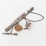 Punhal miniatura com broche e pendentes confeccionado em prata, estrutura posterior composta por alfinete articulado e fecho tipo gancho. Na face inferior, três argolas sustentam correntes em malha fina, das quais pendem miniaturas representando bule, porongo (cuia) em material orgânico em cabaça natural e bomba para chimarrão. Conjunto de caráter temático regional, executado em prata do toque 0,800, conforme marcação observada na peça relacionada do mesmo conjunto, acompanha de punção com as iniciais R.C. Produção sul-americana, final do século XIX / início do século XX. Medida: Comp.: 6,3 cm.