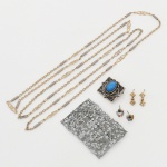 Par de brincos em metal com strass coloridos, dotados de composição frontal em forma circular aberta, estruturada por pedra central facetada, circundada por pedras lapidadas menores em tonalidades diversas (azul, verde, rosa e âmbar), fixadas por garras.Estrutura em metal dourado, com acabamento polido. Pedras em material vítreo (strass), lapidação facetada, cravação em garras. Sistema de fixação por pino reto, destinados a uso com tarraxa. Apresentam marcas de uso e do tempo. Medida: Diam: 1 cm. b) Par de brincos articulados com elementos geométricos, compostos por dois módulos geométricos em forma de poliedros vazados, articulados entre si por argola. Estrutura em metal dourado, com aplicação de pequenas pedras incolores lapidadas, fixadas no interior das faces triangulares por cravação. Sistema de fixação por pino reto. Medida: Comp.: 1,5 cm. c) Broche em metal prateado com pedra azul, formato retangular, dotada de composição central com pedra oval cabochão de coloração azul em material sintético, engastada em bisel e circundada por fileira de granulação metálica. Campo decorado por elementos geométricos e vazados, formando motivo estrelado sobre base retangular com relevos circulares e ornatos em baixo-relevo. Verso com sistema de fecho por alfinete horizontal articulado. Medidas: Alt.: 2,5 x Comp.: 3 cm; d) Broche em metal prateado com marcassitas, formato retangular, estrutura vazada composta por volutas e elementos fitomórficos estilizados. Superfície frontal integralmente aplicada com pedras incolores lapidadas(marcassitas), fixadas por cravação individual. Verso com sistema de fecho por alfinete horizontal articulado. Medidas: Alt.: 4 x Comp.: 6 cm. e) Colar em metal prateado e dourado(com desgaste) com elementos geométricos vazados, composto por segmentos alternados de corrente de elos ovais e elementos intermediários tubulares lisos, intercalados por módulos vazados de desenho geométrico em forma losangular. Medida: Comp.: 60 cm.