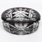 Cinzeiro circular em cristal lapidado, apresentando corpo espesso com sobreposição cromática em tom ametista, posteriormente lapidada para revelar o cristal incolor inferior, criando contraste decorativo; aba superior apresentando quatro descansos para cigarro moldado com reentrâncias semicirculares, corpo externo ornamentado por lapidação facetada em motivos geométricos e radiais, destacando-se composição em forma de estrela estilizada nas paredes laterais; fundo interno apresenta lapidação estrelada(star-cut), destinada a intensificar a refração da luz. Medidas: Alt.: 5 x Diam.: 15 cm.