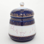 Biscoiteira em porcelana esmaltada, de corpo cilíndrico ligeiramente bojudo, com ombro arredondado e boca circular. Decoração composta por largas faixas em azul cobalto com aplicação de motivos florais e arabescos em douração, dispostos em frisos contínuos. A zona central apresenta campo em esmalte branco ornamentado por pequenos motivos florais estilizados em policromia rosada, distribuídos de maneira esparsa. Tampa ligeiramente abaulada, igualmente esmaltada em azul cobalto com rica douração floral, rematada por puxador em forma de botão esférico em branco, base plana com anel de assentamento. Marca na base em carimbo rosado: CERÂMICA SÃO JOSÉ LTDA.  PEDREIRA, manufatura brasileira do polo cerâmico de Pedreira, São Paulo. Medidas: Alt.: 22,5 x Diam.: 15 cm. Apresenta desgastes na douradura, vide foto.