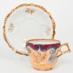 Belíssima xícara para chá em porcelana esmaltada, corpo campaniforme e pires circular;  apresenta bojo facetado com decoração em relevo de motivo vegetalista e ornamental, composta por volutas, folhas estilizadas e elementos florais modelados, com aplicação de douração abundante. A ornamentação é complementada por faixas e campos em esmaltação policroma, destacando tons de vermelho vinoso, azul e lilás, delimitados por filetes e contornos em dourado;  asa lateral estilizada igualmente realçada por douração, borda superior marcada por filete dourado contínuo, pires apresenta aba ondulada com borda recortada e dourada, campo interno branco com reservas decoradas por pequenos ramalhetes florais dourados, distribuídos radialmente e separados por sulcos moldados. Medidas: Xícara: Alt.: 7,5 x Comp.: 12 x Larg.: 9 cm; Pires: Diam.: 15 cm.