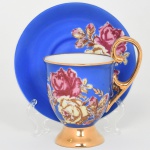 Porcelanarte, Campo Largo - Paraná. Belíssima chávena para chá em porcelana esmaltada, bojo campaniforme elevado sobre pequeno pé circular, acompanhada de pires circular de aba larga, decoração apresenta fundo em azul cobalto intenso, ornado por ramalhetes de rosas estilizadas em policromia, predominando tons de rosa, creme e marrom, dispostos lateralmente tanto na xícara quanto no pires,  xícara possui borda e pé inteiramente dourados, bem como asa em voluta com aplicação integral de douração, interior permanece em esmalte branco com filete dourado junto à borda, pires apresenta campo central em azul cobalto com reserva decorada por composição floral correspondente. Marca na base da xícara em verde, composta por monograma P entre ramos de louro sobre a inscrição PORCELANARTE  PARANÁ, manufatura brasileira. Medidas: Chávena: Alt.: 12 x Comp.: 11 x Larg.: 8 cm; Pires: Diam.: 15 cm.