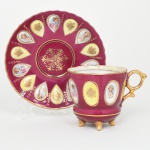 Exuberante chávena para chá executada em porcelana japonesa, bojo cilíndrico ligeiramente campaniforme, elevada sobre três pés terminados em voluta, acompanhada de pires circular de aba recortada. Decoração em fundo de tom rosáceo com ampla aplicação de douração, apresentando reservas em forma de medalhões ovais e em gota distribuídas ao redor da peça, contendo miniaturas pintadas representando bustos femininos em trajes de época, alternadas com campos em esmalte amarelo ornados por motivo floral em dourado; superfície enriquecida por filetes, pontilhados e frisos dourados em relevo.; chávena apresenta asa lateral vazada de desenho ornamental, inteiramente dourada, pires repete o esquema decorativo da chávena, com sequência radial de reservas contendo miniaturas e motivo floral, centradas por ornato vegetalista estilizado em douração ao centro. Marca da manufatura no verso em dourado: MARCA REGISTRADA  K.K.R.  MADE IN JAPAN. Medidas: Chávena: Alt.: 8,5 x Comp.: 11,5 x Larg.: 8,5 cm; Pires: Diam.: 15 cm.