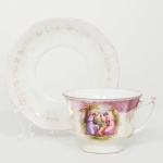 Xícara para chá  em porcelana esmaltada, bojo campaniforme, facetado com oito lados, assente sobre pé baixo, acompanhada de pires octogonal com borda suavemente recortada. Decoração em esmaltação predominantemente branca com faixa superior em tom rosáceo, enriquecida por ornatos florais e filetes em douração. No bojo da xícara apresenta-se reserva circular com cena figurativa policroma, representando duas figuras femininas em trajes clássicos junto a um putto em ambiente campestre, circundada por moldura ornamental dourada, pires apresenta decoração harmonizada com a xícara, composta por filetes e delicados motivos florais dourados e rosados dispostos ao longo da aba. A xícara possui asa lateral de perfil curvo, parcialmente realçada por douração. Apresenta acentuado desgaste na douradura e na policromia, vide foto. Ausência de marca de manufatura. Medidas: Alt.: 6 x Comp.: 10,5 x Larg.: 9 cm; Pires: Diam.: 13,5 cm.