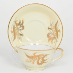 Xícara para chá em porcelana esmaltada, bojo campaniforme apoiado sobre pequeno pé anelar, acompanhada de pires circular; superfície apresenta fundo em tonalidade marfim com suave matização amarelada, enriquecida por decoração estilizada em douração representando espigas de trigo e folhas alongadas, dispostas de forma fluida e assimétrica ao redor do bojo e da aba do pires, borda da xícara e do pires delimitada por filete dourado contínuo, asa lateral em voluta realçada por douração. Marca da manufatura no verso acompanhada da inscrição YOSHINO. Medidas: Xícara: Alt.: 5,5 x Comp.: 12 x Larg.: 9,5 cm; Pires: Diam.: 14 cm.