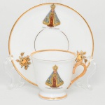 Porcellana Mauá, Mauá - SP(Fundada em 1937 por Fritz Erwin Schmidt / ativa até 1968). Xícara para café em porcelana nacional, bojo campaniforme apoiado sobre pequeno pé anelar e pires circular, decoração apresenta fundo branco com filetes dourados nas bordas, bem como asa lateral em voluta  inteiramente dourada; bojo da xícara e o pires apresentam imagem em estampagem policromada de Nossa Senhora Aparecida, representada com coroa e manto azul ricamente ornamentado em dourado, acompanhada da inscrição Lembrança de N. Sra. Aparecida, característica de peças devocionais e de recordação do santuário, pires complementado por ramalhetes florais estilizados em douração distribuídos ao redor da aba. Marca da manufatura no verso. Medidas: Xícara: Alt.: 5 x Comp.: 7 x Larg.: 5,5 cm; Pires: Diam.: 10,5 cm. Apresenta leve desgaste na inscrição do pires.