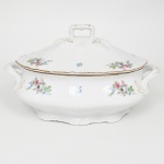 Count Thun Porcelain Factory, Kláterec - República Tcheca(Fundada em 1794). Sopeira em porcelana esmaltada, de corpo ovalado e bojo abaulado, apoiada sobre base anelar recortada. Apresenta duas alças laterais integradas, de perfil curvo e vazado, acompanhando a linha do recipiente; tampa de perfil abaulado e borda ondulada, rematada por puxador central moldado em forma de alça vazada, com delicados acentos em douração. Possui ainda recorte lateral (entaille) destinado à passagem da concha de serviço. Decoração apresenta fundo branco com pequenos ramalhetes florais policromos distribuídos de maneira regular pelo corpo e pela tampa, compostos por flores em tons de cinza, rosa, azul e verde; ornamentação complementada por motivos estilizados em douração nas alças, bordas e relevos da tampa, além de filete dourado acompanhando o contorno ondulado da borda. Marca na base em verde: coroa sobre as iniciais TK e a inscrição Thun, manufatura da porcelana Thun, tradicional fábrica da Boêmia (atual República Tcheca). Medidas: Alt.: 18,5 x Comp.: 31,5 x Larg.: 23 cm. Apresenta pequena quebradura na borda da base, demanda pequeno restauro.