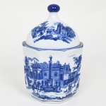 Bombonière em porcelana azul-e-branco de inspiração Delft, corpo facetado, acompanhado de tampa abobadada encimada por pega elipsoide; decoração executada em azul monocromático sob esmalte, representando cenas paisagísticas com arquitetura, figuras humanas e elementos campestres, inspiradas na tradição decorativa das faianças holandesas de Delft, corpo apresenta painéis figurativos envolvendo edificações, colunatas e personagens em ambiente urbano e rural, enquanto a tampa repete a temática paisagística, borda realçada por filete azul. Peça decorativa ou utilitária destinada ao serviço de mesa, podendo funcionar como açucareiro ou bombonière. Medidas: Alt.: 20 x Diam.: 13 cm.