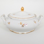 Porcelana Rio Branco/Cia. Brasileira de Vidros, Rio de Janeiro - RJ(Fundada em 1947, ativa até 1967). Terrina com tampa em porcelana nacional da década de 1950; esmaltagem vítrea na cor branca, dotada de decoração em estampagem policromada, com padrão de flores predominando as tonalidades rosa, amarelo, castanho e cinza, realçado com generosos arremates de frisos em douradura, tampa e bojo com leve movimento ondulatório modelado na própria pasta, alças laterais reviradas para cima, puxador ovoide. Marca da manufatura no verso. Medidas: Alt.: 15,5 x Comp.: 27 x Larg.: 22 cm. Apresenta quebradura na borda da boca.