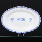 ROSSLAU - ALEMANHA. Exuberante e grande travessa oval em porcelana alemã, de contorno recortado com aba larga levemente ondulada. Esmaltação branca, decoração apresenta composição em azul padrão china-blau, característica da tradição decorativa centro-europeia inspirada nas porcelanas orientais; aba ornada por friso contínuo em azul com motivos florais estilizados e elementos geométricos, delimitado por filete dourado acompanhando o contorno ondulado da borda, campo central apresenta vinheta decorativa em azul, composta por arranjo floral acompanhado de pequeno elemento ornamental representando ânfora sobre base. Marca na base em azul: CHINA-BLAU / ROSSLAU, indicando manufatura da fábrica Rosslau, Alemanha. Medidas: Comp.: 41,5 x Larg.: 28,5 cm.
