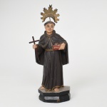 ARTE SACRA. Santo Antônio. Imagem sacra executada em madeira entalhada e policromada. Apresenta de pé sobre base prismática escalonada, trajando hábito franciscano de mangas largas e capuz posterior, cingido por cordão à cintura, com pés descalços apoiados sobre pequeno plinto, sustenta na mão direita cruz latina e na esquerda livro onde se assente o Menino Jesus(ausente neste exemplar), atributo iconográfico associado ao santo pregador franciscano; cabeça encimada por resplendor metálico dourado em forma de raios, fixado por haste posterior; superfície apresenta policromia em tons escuros no hábito, com filetes dourados delimitando pregas e bordas do vestuário, e tratamento naturalista do rosto e das mãos. Medidas: Alt.: 20,5 x Comp.: 8 x Larg.: 5 cm. Apresenta perda do Menino Jesus, e pequenos desgastes visíveis nas fotos.