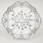 Carl Tielsch, Silésia - Alemanha. Travessa em porcelana alemã, comemorativa de bodas de prata; esmaltação branca, de contorno lobado com bordas recortadas e vazadas, dotada de pequenas pegas laterais integradas, aba apresenta ornamentação em prata aplicada, composta por volutas, elementos vegetalistas estilizados e reservas vazadas, formando composição decorativa contínua de inspiração rocaille, campo central ornado por ramagens florais estilizadas em prata, envolvendo inscrição caligráfica em alemão executada em escrita gótica: "Seiner Silberhochzeit zum Tage den 28 Jahr". Marca da manufatura no verso. Medidas: Alt. 5 x Comp.: 31 x Larg.: 29 cm. Apresenta desgaste na escrita do centro, vide foto.