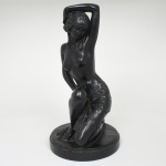 Escultura decorativa representando figura feminina seminua em postura sinuosa, ajoelhada sobre uma das pernas e com o braço elevado sobre a cabeça, composição de forte caráter ornamental inspirada na estética Art Déco, apresenta tratamento estilizado das formas, com ênfase nas linhas curvas do corpo e na elegância do gesto, é representada com cabelos curtos ondulados e adorno de inspiração orientalista, usando apenas tecido drapeado envolvendo a região dos quadris, decorado com elementos geométricos e relevos que evocam motivos decorativos característicos do período entre-guerras. A superfície recebe acabamento em pátina de tonalidade preta uniforme, simulando bronze, assentada sobre base circular integrada, originalmente destinada à montagem como base de luminária ou elemento decorativo, evidenciada pelos orifícios presentes na base e no corpo da peça, destinados à passagem de fiação elétrica. Medidas: Alt.: 35 x Diam.: 18 cm. Apresenta pequenos desgastes na pátina em pontos isolados.