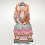 Escultura de bodhisattva Guanyin em porclelana policroma esmaltada, manifestação chinesa do bodhisattva Avalokitevara, divindade central do budismo mahayana associada à compaixão universal. Apreseta-se representada sentada em posição de meditação (padmsana) sobre grande flor de lótus aberta, motivo iconográfico que simboliza pureza espiritual, iluminação e transcendência do mundo material. A divindade apresenta expressão serena e contemplativa, com olhos semicerrados e traços faciais suavizados. Os cabelos são modelados em pequenas volutas azuis, encimados pela característica protuberância craniana (ua) adornada por elemento central, ao redor da cabeça observa-se auréola circular com contorno flamejante, ricamente ornamentada com colares e adornos peitorais, característica típica das representações de bodhisattvas, que diferem das representações do Buddha histórico por apresentarem aparência principesca e adornada; apresenta esmaltação vítrea policroma aplicada após a modelagem, com acabamento brilhante e riqueza cromática típica de esculturas devocionais chinesas produzidas para altares domésticos ou pequenos santuários. Apresenta quebradura nas mãos e na auréola, demanda restauro. Medidas: Alt.: 32 x Comp.: 16 x Larg.: 12 cm.