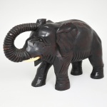 Escultura animalista representando elefante, esculpida em bloco de madeira, com acabamento polido e tonalidade escura. Figura em posição de marcha, com tromba elevada e curvada, apresentando sulcos rítmicos ao longo da extensão, presas aplicadas em material sintético de coloração clara e olhos entalhados. Corpo com tratamento volumétrico simplificado, marcado por incisões lineares que sugerem textura da pele e definição anatômica das orelhas, membros e dorso. Medidas: Alt.: 23 x Comp.: 37 x Larg.: 12 cm.