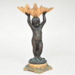 Excepcional escultura francesa executada em bronze, representando putto, modelado em posição frontal, braços elevados sustentando grande concha naturalística disposta horizontalmente; superfície apresenta pátina escura de tonalidade castanho-bronzeada, com áreas de oxidação esverdeada e realces em douração na concha e em elementos ornamentais da base. Apresenta-se de pé, com leve flexão de uma das pernas, apoiada sobre base circular ornada em relevo com elementos de temática marinha, incluindo conchas e vegetação aquática estilizada, base assenta sobre plinto moldurado de contorno recortado, decorado por volutas e folhas de acanto, com vestígios de douração. A concha superior apresenta modelagem naturalista de bordas onduladas e superfície irregular, configurando receptáculo aberto, concebido como elemento funcional e decorativo. Estado conforme fotografias. Medidas: Alt.: 30 x Comp.: 16,5 x Larg.: 13,5 cm.