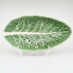 Travessa oval executada em cerâmica modelada em formato de folha, de contorno alongado e borda elevada, interior côncavo apresentando nervura central em relevo e ramificações laterais estilizadas. Superfície esmaltada em tonalidade verde com variações cromáticas, com as nervuras ressaltadas em branco, configurando efeito naturalista. Base oval com marca em relevo NARA FP. Medidas: Alt.: 7 x Comp.: 39,5 x Larg.: 20 cm.