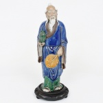 Estatueta de sábio oriental em cerâmica chinesa esmaltada policroma, relacionada à tradição cerâmica de Shiwan. Figura escultórica representando ancião oriental de feições serenas, com cabeça parcialmente calva, longas sobrancelhas e barba fluente, trajando amplas vestes tradicionais, segura pequeno vaso na mão diireita e leque na mão esquerda, atributos associados a personagens da tradição filosófica e simbólica chinesa, frequentemente vinculados à ideia de sabedoria, contemplação e longevidade, sendo tipologicamente relacionada às representações do imortal taoista Shou Xing, divindade da longevidade na cultura chinesa. A modelagem apresenta tratamento expressivo do rosto e das vestes, com pregas bem definidas e cuidadoso detalhamento das feições. A peça é revestida por esmaltação vítrea, predominando azul intenso nas vestes, complementado por tons verdes na parte inferior, marrom no forro das mangas, amarelo no leque e esmalte claro na barba e cabelos, combinação cromática característica das figuras inspiradas na tradição cerâmica de Shiwan, conhecida pela produção de esculturas de sábios, imortais e personagens da cultura chinesa.Base circular em madeira entalhada. Marcada ao fundo  CHINA, indicando produção chinesa destinada à exportação. Medidas: Alt.: 29 x Comp.: 14 x Larg.: 11 cm.