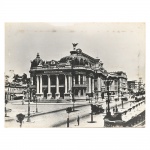 Fotografia em preto e branco, impressão sobre papel aplicada sobre moldura de madeira, representando o edifício do Theatro Municipal do Rio de Janeiro, com fachada de composição eclética, marcada por colunata frontal, cúpulas laterais e central encimada por escultura alegórica. Cena urbana com circulação de pedestres, veículos automotores de época e postes de iluminação pública distribuídos ao longo da via. Medidas: Alt.: 29 x Comp.: 39 cm.