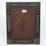 Moldura porta-retrato em madeira entalhada com aplicações em prata, formato retangular com moldura talhado em tremidos. Montantes laterais com colunas em relevo, decoradas por motivos florais em repetição vertical, cantos reforçados com blocos quadrangulares entalhados com rosetas em relevo. Verso com tampa móvel articulada e apoio posterior basculante. Presença de etiqueta de comerciante com inscrição J. J. Barbosa & Cia., R. Senador da Silva, 63  Catete  Rio  Indústria Brasileira. Medidas: Alt.: x Comp.: