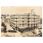 Fotografia em preto e branco aplicada sobre moldura de madeira, impressão sobre papel, representando o Hotel Avenida, no Rio de Janeiro, edifício de esquina em área urbana com circulação de bondes e transeuntes ao longo das vias adjacentes. Medidas: Alt.: 29 x Comp.: 39 cm.