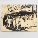 Fotografia em preto e branco aplicada sobre moldura de madeira, impressão sobre papel, representando bonde urbano com passageiros embarcados e grupo de transeuntes ao redor, em via ladeada por edificações com balcões e vãos em série, e placa publicitária Beba Caxambu, da marca de água mineral Caxambu, posicionada na lateral superior do bonde. Medidas: Alt.: 29 x Comp.: 39 cm.