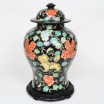 Potiche com tampa em porcelana chinesa decorada em esmaltes policromos sobre fundo de tonalidade preta, portando marca apócrifa Qianlong, China, século XX. Corpo balaústre com ombro largo e gargalo curto, acompanhado de tampa abaulada com botão central, executado em porcelana e apoiado sobre base circular entalhada em madeira, provavelmente posterior, superfície de fundo preto vidrado, sobre o qual se desenvolve decoração pintada em esmaltes policromos em tons de verde, vermelho-alaranjado, amarelo, azul e branco, ornamentação composta por grandes peônias estilizadas e ramos vegetalistas sinuosos, com folhas em verde-jade que se espalham de forma contínua por toda a superfície do vaso; entre os elementos florais aparecem leões guardiões chineses (shishi ou cães de Fó) representados em diferentes cores, inseridos entre os ramos decorativos. As peônias, motivo tradicional da porcelana chinesa, são associadas à prosperidade, honra e riqueza, enquanto os leões guardiões simbolizam proteção, força e poder auspicioso. A decoração se estende também à tampa, mantendo o mesmo repertório floral e cromático. Apresenta ao fundo marca em selo vermelho com quatro caracteres chineses - (Qianlong nian zhi), traduzido como feito durante o reinado de Qianlong, referência ao imperador Qianlong Emperor, da Qing Dynasty. Trata-se, contudo, de marca de homenagem ou apócrifa, amplamente utilizada em porcelanas decorativas chinesas posteriores. Medidas: Alt.: 43,5 x Diam.: 28 cm.