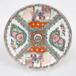 Prato medalhão em porcelana chinesa decorado em esmaltes policromos no estilo famille-rose, esmaltação branca, com decoração executada em esmaltes policromos sobre o vidrado, predominando tons de rosa, verde, vermelho-ferro, azul e amarelo, com contornos definidos por traço escuro e filete dourado no bordo, composição decorativa organiza-se em quatro reservas principais dispostas radialmente, alternando cenas figurativas e arranjos florais estilizados. Duas reservas apresentam cenas de interior com personagens chineses trajando vestes tradicionais, representados em ambiente arquitetônico com balaustrada ao fundo, mesa e elementos decorativos, típicos da iconografia doméstica da porcelana chinesa exportada, as outras reservas apresentam arranjos florais compostos por peônias, folhas estilizadas e pequenos pássaros, elementos simbólicos recorrentes na porcelana chinesa, associados à prosperidade e à felicidade,  centro com medalhão circular com pequeno motivo floral, circundado por ornamentação vegetal e geométrica, aba delimitada por molduras ornamentais sinuosas em verde e rosa, com elementos rocaille estilizados que criam a divisão entre as reservas. Verso apresenta marca em selo vermelho com caracteres chineses, aplicada sob o vidrado, comum em porcelanas chinesas produzidas para exportação no século XX, inspiradas em modelos decorativos do período Qing. Medida: Diam.: 30,5 cm.