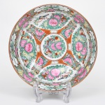 Grande tigela executada em porcelana chinesa decorada em esmaltes policromos no estilo famille-rose; esmaltação branca, formato hemisférico profundo, apoiada sobre anel de base circular, com decoração realizada em esmaltes policromos sobre o vidrado, predominando tons de rosa, verde, vermelho-ferro, amarelo e azul, com contornos definidos por traço escuro; interior apresenta composição radial organizada em reservas ornamentais, separadas por elementos geométricos e painéis em vermelho-ferro. No centro encontra-se medalhão circular com arranjo floral estilizado, composto por peônias e folhagens, circundado por motivos vegetais, pequenos insetos e borboletas, reservas principais apresentam arranjos florais exuberantes com peônias, frutos e folhagens, acompanhados por aves e borboletas, motivos recorrentes na porcelana chinesa e associados a símbolos de prosperidade, longevidade e felicidade, parede externa repete o mesmo repertório decorativo, alternando cartelas em branco com ramos florais estilizados e painéis com flores, pássaros e elementos vegetalistas, separados por faixas com padrão geométrico inspirado no meandro grego, característica frequentemente presente em porcelanas chinesas de exportação, verso apresenta marca em selo vermelho com caracteres chineses, aplicada sob o vidrado no interior do anel de base, comum em produções decorativas chinesas inspiradas em marcas de reinado da dinastia Qing. Medidas: Alt.: 10 x Diam.: 25,5 cm.