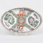 Travessa oval executada em porcelana chinesa decorado em esmaltes policromos no estilo famille-rose, esmaltação branca, com decoração executada em esmaltes policromos sobre o vidrado, predominando tons de rosa, verde, vermelho-ferro, azul e amarelo, com contornos definidos por traço escuro e filete dourado no bordo, composição decorativa organiza-se em quatro reservas principais dispostas radialmente, alternando cenas figurativas e arranjos florais estilizados. Duas reservas apresentam cenas de interior com personagens chineses trajando vestes tradicionais, representados em ambiente arquitetônico com balaustrada ao fundo, mesa e elementos decorativos, típicos da iconografia doméstica da porcelana chinesa exportada, as outras reservas apresentam arranjos florais compostos por peônias, folhas estilizadas e pequenos pássaros, elementos simbólicos recorrentes na porcelana chinesa, associados à prosperidade e à felicidade,  centro com medalhão circular com pequeno motivo floral, circundado por ornamentação vegetal e geométrica, aba delimitada por molduras ornamentais sinuosas em verde e rosa, com elementos rocaille estilizados que criam a divisão entre as reservas. Verso apresenta marca em selo vermelho com caracteres chineses, aplicada sob o vidrado, comum em porcelanas chinesas produzidas para exportação no século XX, inspiradas em modelos decorativos do período Qing. Medida: Alt.: 3,5 x Comp. 35 x Larg.: 23 cm.
