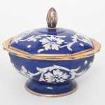 Cerâmica Conrado Bonadio, São José dos Campos - SP (Fundada em março de 1936 por Corado Bonadio). Bombonière com tampa em cerâmica esmaltada. Bojo hemisférico facetado com oito lados, assente sobre base circular levemente elevada. Corpo e tampa executados em porcelana esmaltada em fundo azul cobalto, decorados com composição floral estilizada em esmalte branco, acompanhada de filetes e contornos em douração aplicada. Tampa de perfil abaulado, acompanhando a geometria octogonal do corpo, encimada por pomo em forma de botão alongado, igualmente decorado com douração. Bordas onduladas realçadas por faixa dourada contínua. Interior esmaltado em branco. Base apresentando inscrição manuscrita Bonadio, além de numeração 143 2C aplicada em esmalte. Medidas: Alt.: 17 x Diam.: 20,5 cm. Tampa apresenta acentuada rachadura, e leves desgastes na douradura.