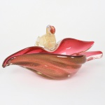 Centro de mesa em forma de folhagem em vidro de Murano. Corpo em forma orgânica com bordas onduladas e abas assimétricas. Vidro translúcido em tonalidade rosa com generosa inclusão de pó de ouro, associado a aplicação escultórica representando ave em vidro incolor com tonalidade dourada, modelada a quente, com olho em vidro aplicado. Atribuído a ARQUIMEDES SEGUSO. Apresenta acentuadas quebraduras, demanda restauros. Medidas: